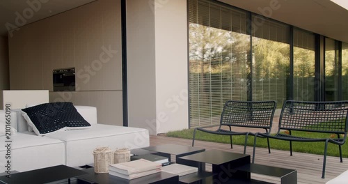 Modern home backyard patio area featuring white sofa, black wicker chairs, black square tables, wood deck, grass lawn, and glass sliding doors with blinds and trees reflecting in the glass.