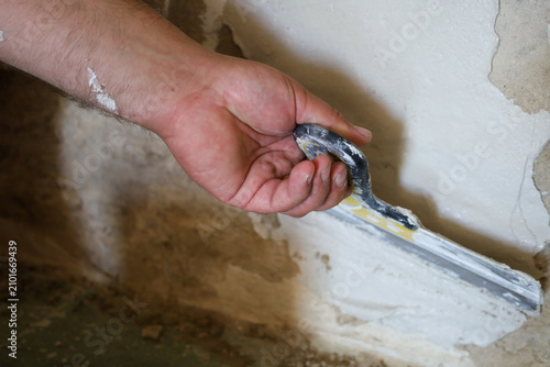 Fotografie A worker's hand firmly holds a construction spatula, applying white plaster to an old uneven wall during renovation