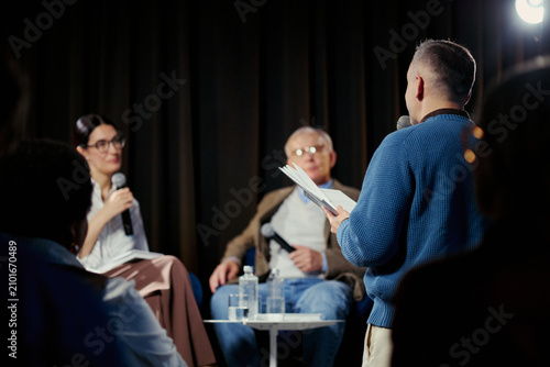 Obraz na płótnie Young adult man speaking into microphone and reading notes during panel discussion, engaging speakers on stage