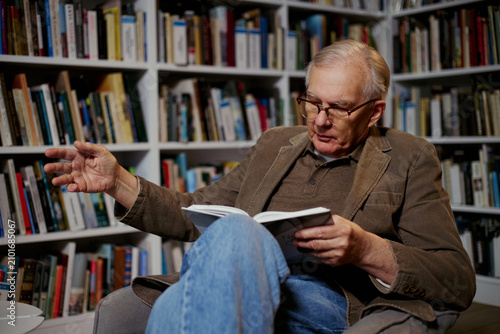 Fotografie Senior Caucasian man reading book and gesturing while explaining ideas in home library, conveying expertise and lifelong learning