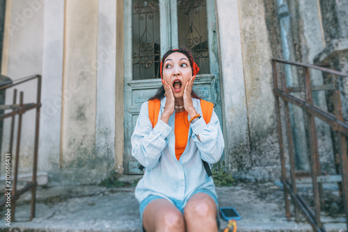 Фотография Shocked young woman traveler with hands on face sitting on steps of an abandoned