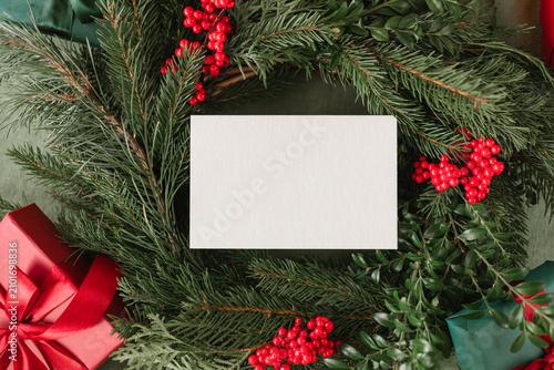 A blank white paper card, framed by a lush Christmas wreath with red berries, and wrapped gift boxes with satin bows.
