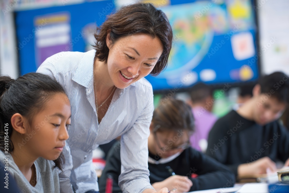 Fototapeta premium back to school - caring teacher of asian descent kneeling or leaning next to a student's desk, helping with a difficult assignment. asian student looks focused, appreciative.