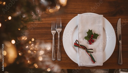A festive Christmas dinner table setting with a white plate, silverware, and a napkin adorned with holly and berries, surrounded by bokeh lights