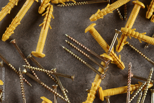 Yellow plastic dowels with metal screws on a gray background. A pile of yellow quick-installation construction dowels and metal self-tapping screws scattered on a gray textured surface. Fasteners