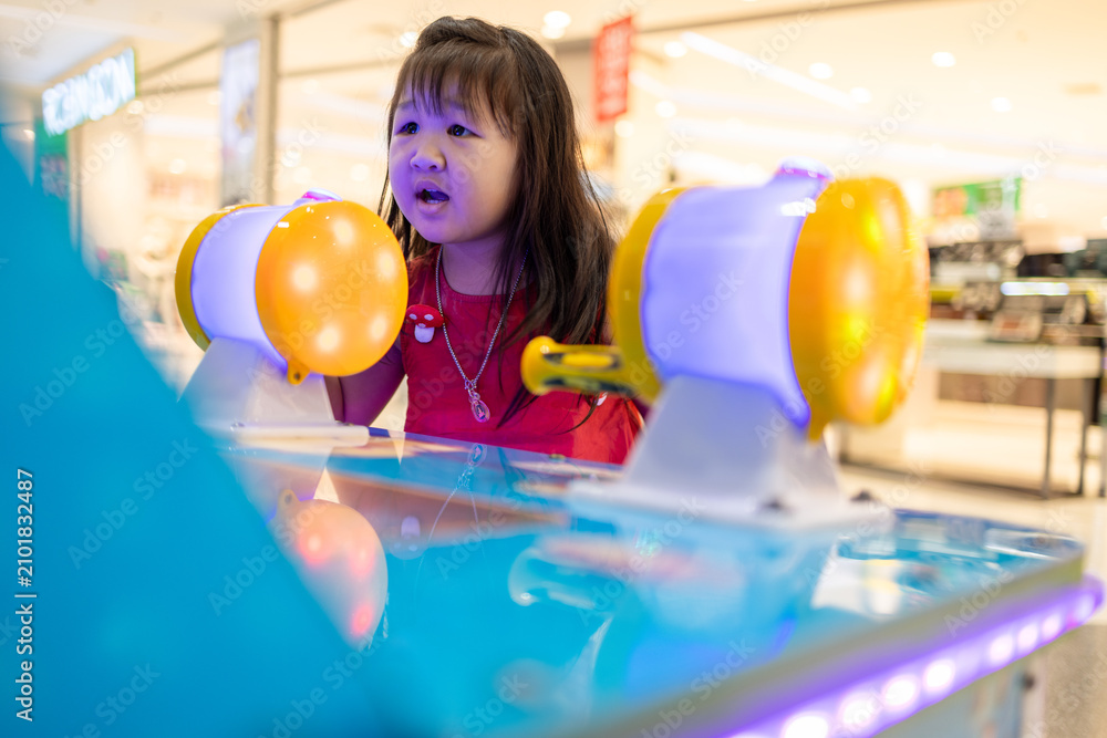 Fototapeta premium Happy Little Girl Playing Arcade Game at Shopping Mall,A cute Asian little girl having fun and playing an interactive electronic arcade game at an amusement center inside a shopping mall during.