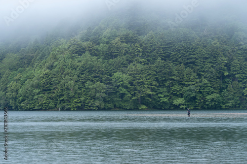 梅雨の雨が降る湯ノ湖と深緑の山々と釣り人