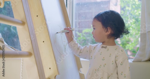 A focused toddler with a hearing aid practices drawing on a whiteboard easel, engaging in creative play and early childhood development at home in a sunlit room.