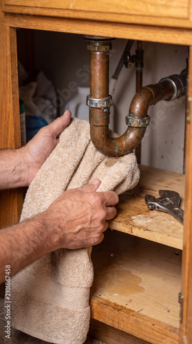 Close-Up Of Hands Fixing Small Leaking Pipe Under Kitchen Sink With Towel And Wrench, Home Repair Concept
