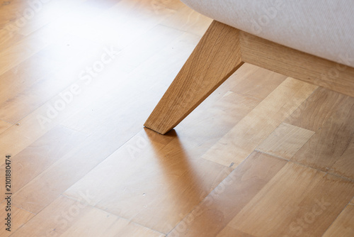 A detailed close up shot of a modern wooden sofa leg resting on a bright polished hardwood parquet floor in a home interior.