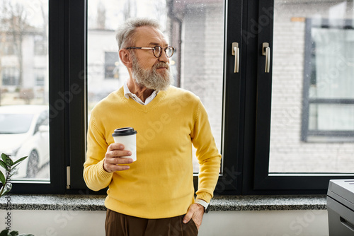 Canvas Print Mature man with a beard holds a coffee cup while gazing thoughtfully out the window