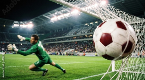 Professional goalkeeper in green uniform diving to save a soccer ball flying into the net during an intense night match at a large stadium with bright artificial floodlight illumination visible