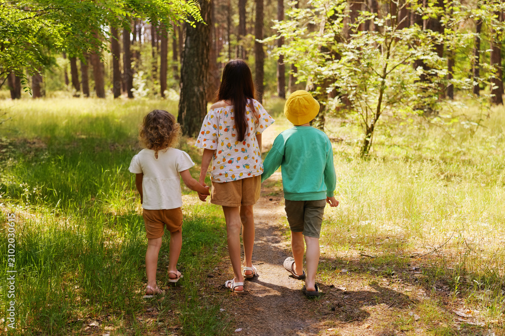 Fototapeta premium Three children stroll down a dirt path in a forest. They hold hands and enjoy the sunny day surrounded by trees and plants