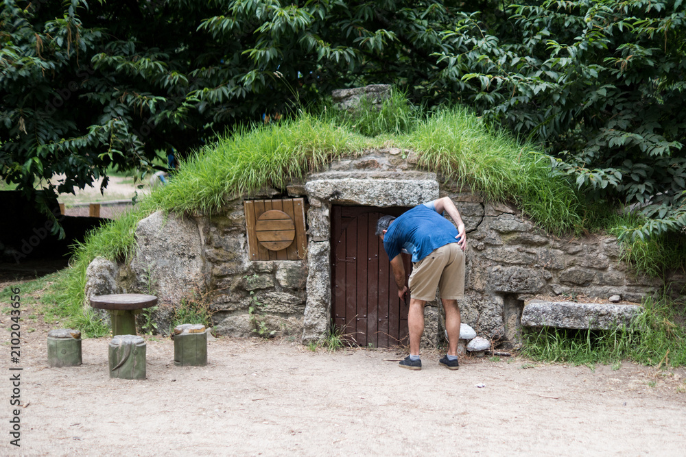 Fototapeta premium A male tourist bends down to explore the miniature wooden door of a whimsical stone earth house with a grass roof during a family summer vacation.