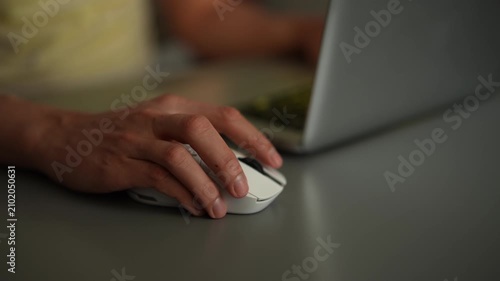 Close-up video of persons hand using white wireless mouse. User clicks and scrolls working on laptop in dark room. Screen light reflects on desk, creating dynamic mood. Shooting in slow motion.