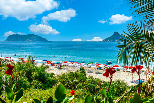 Beautiful tropical beach of Barra da Tijuca in Rio de Janeiro, Brazil. Colorful umbrellas on white sand, vibrant blue ocean, islands, and lush foliage under a sunny sky.