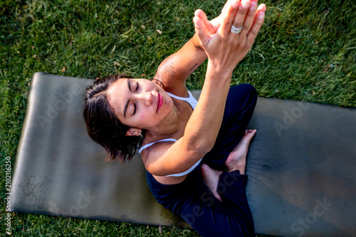 Obraz na plátně A young Hispanic woman practises a reclining stretch on a yoga mat in a lush green park at golden hour, a practice of mindfulness and mental wellbeing in the tranquil evening
