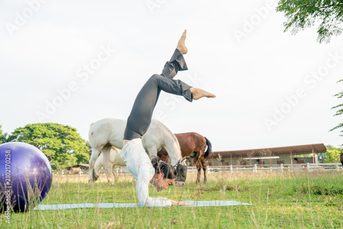 Active Asian woman practicing advanced yoga poses on a mat at a scenic horse farm meadow.