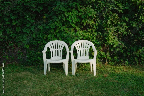 Two white plastic chairs sit on a grassy lawn before a wall covered in lush green vines.  The scene is tranquil and subtly evokes a sense of waiting or anticipation