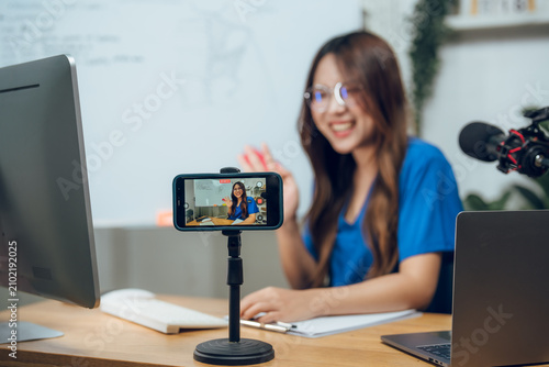 Cheerful Asian female nurse with glasses recording health vlog on smartphone in modern clinic. Young medical professional broadcasting online anatomy lesson and virtual care telemedicine tutorial.