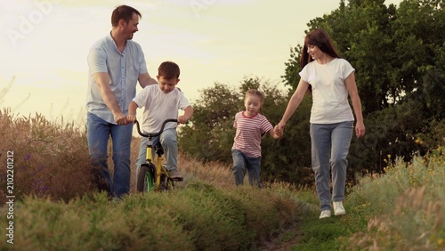 Bild auf Leinwand Mother holds running girl and father supports boy riding bicycle on family vacation at country area