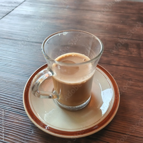 Hot Milk Coffee in Clear Glass Cup on Ceramic Saucer over Wooden Table, Cozy Cafe Beverage Close Up