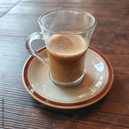 Hot Milk Coffee in Clear Glass Cup on Ceramic Saucer over Wooden Table, Cozy Cafe Beverage Close Up