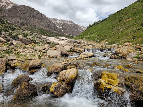 A rocky mountain stream cascades over moss-covered boulders, with patches of avalanche snow and rugged snow-capped peaks rising behind green hillsides.
📍 Hawraman Takht, Kurdistan, Iran 