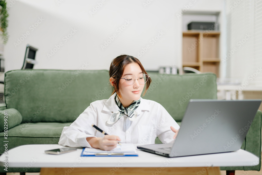 Fototapeta premium Asian woman manages business calls on her smartphone while working on a laptop with documents at home, blending corporate flair with cozy remote work.