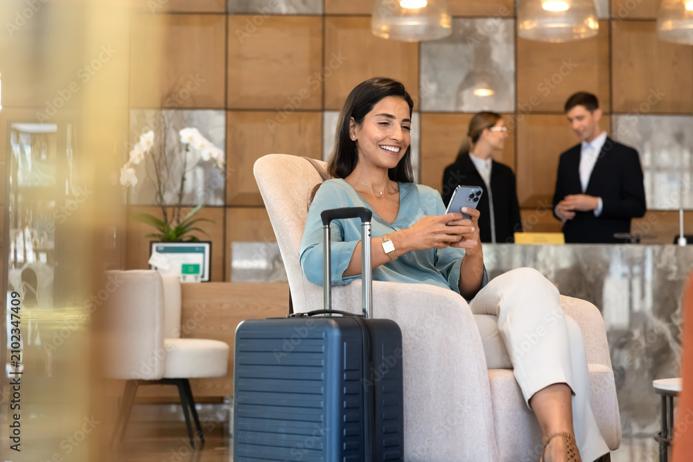 Obraz premium Female traveler guest sitting in hotel lobby, checking smartphone to read messages, confirm booking, browse travel information, using online customer services. Digital communication, hotel industry