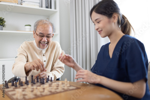 Smiling senior Asian man playing chess with female nurse caregiver..Elderly man in wheelchair playing board game with home healthcare nurseProfessional female caregiver supporting senior patient.