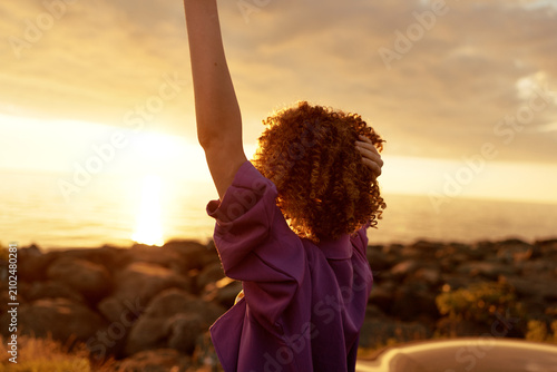 Rear view of a woman with curly hair raising her arm at sunset by the sea  wearing a purple top  capturing a carefree lifestyle moment and warm golden light.