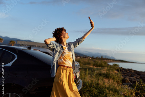 Konstfotografi Cheerful woman outdoors, sunlit moment beside a car, coastal background, relaxed