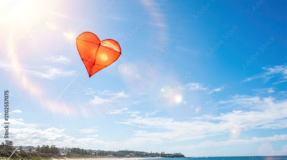 Fototapeta premium Bright red heart-shaped kite flying in sunny beach sky