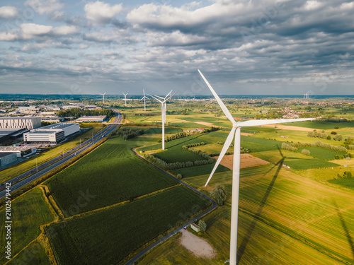 Aerial drone view of wind turbines standing among green patchwork farm fields beside a motorway under a cloudy summer sky, a scene of renewable wind energy and sustainability.