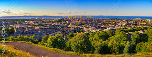 Quadro su tela Panoramic sunset view from Calton Hill over Edinburgh rooftops, Leith Docks, and the Firth of Forth, with green hillside foreground and warm evening light in Scotland, United Kingdom