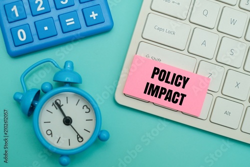 Fotografie Pink sticky note with POLICY IMPACT text on a keyboard, next to a blue alarm clock and calculator against coloured background