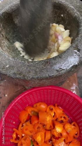 Crushing garlic in a traditional stone mortar and pestle with sliced red chili peppers in a basket foreground.
