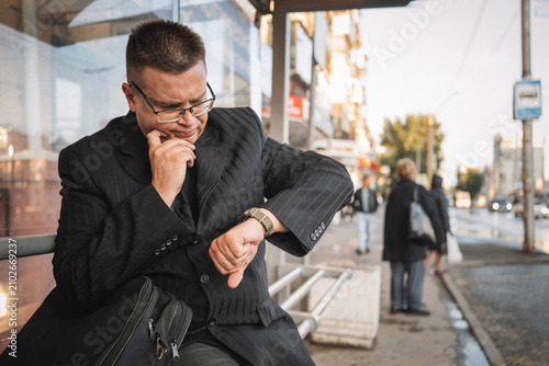 Tablou pe pânză Businessman sitting at a bus stop, looking anxiously at his watch while waiting for transport