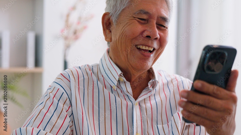 Fototapeta premium Happy senior Asian grandfather using smartphone for video call or watching online content at home. Cheerful elderly man enjoying modern digital technology lifestyle connection.