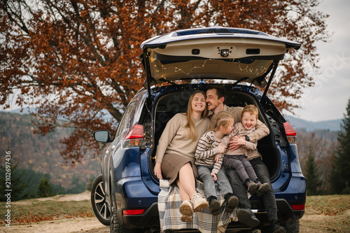 A joyful family, including a father, mother, son, and daughter, sharing a candid, happy moment while sitting in the open trunk of a blue SUV during an autumn mountain trip.