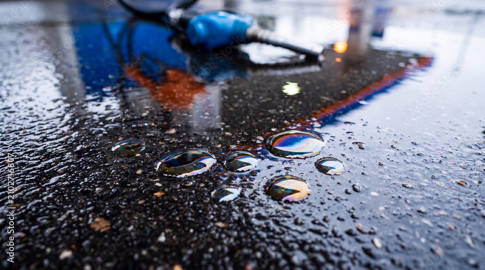 Obraz premium Fuel nozzle on wet asphalt with oil spill creating rainbow sheen, highlighting environmental pollution and energy issues