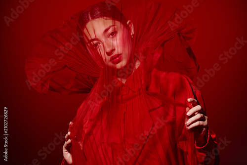 Portrait of a woman in a detailed red fashion dress and veil posing against a red background. Creative beauty concept with dramatic mood and fabric texture.