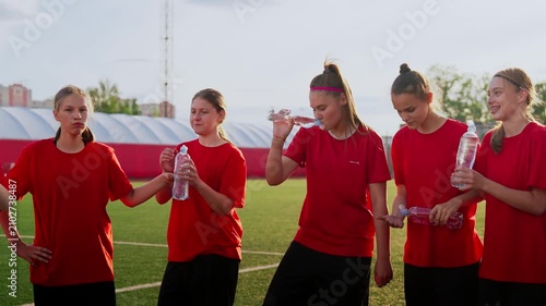 Womens soccer team taking a hydration break during outdoor training session on sunny field, teammates in red jerseys resting and drinking water together