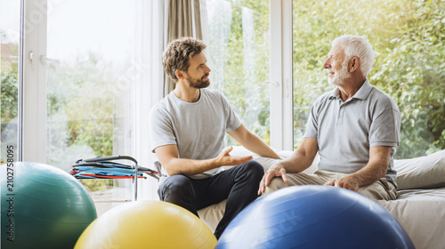 Ultra realistic rehabilitation consultation at home, physical therapist explaining a recovery program to an older man seated comfortably beside a large window