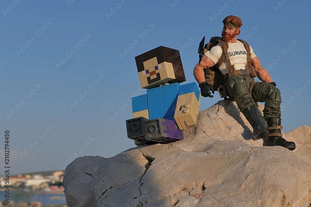 Obraz premium Action figures of Minecraft hero Steve and GI JOE Classified series survival instructor Outback sitting on coastal rock in Croatia, sunlit by evening sunshine, blue skies in background. 