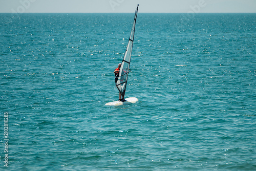 a lone windsurfer riding a white board with a tall sail across a vast, sparkling blue ocean under a clear sky.