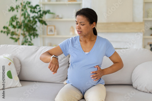 Canvastavla Black pregnant woman checking her smartwatch while holding her belly on sofa, mo