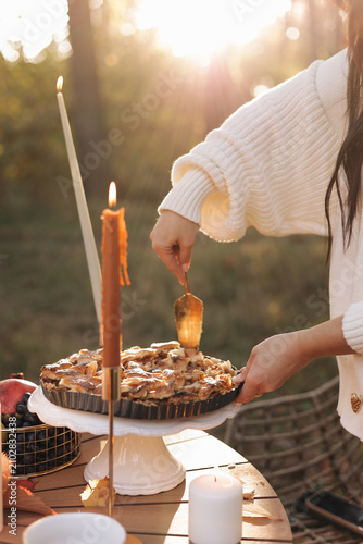A woman in a cozy sweater serves a rustic apple pie on a pedestal during an autumn outdoor picnic, surrounded by candlelight, warm sunlight and seasonal fruit for a relaxed harvest mood.