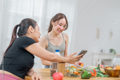 Woman, friend, smartphone, kitchen, salad, vegetable smiling young adult woman and friend looking at smartphone during healthy meal preparation in bright home kitchen